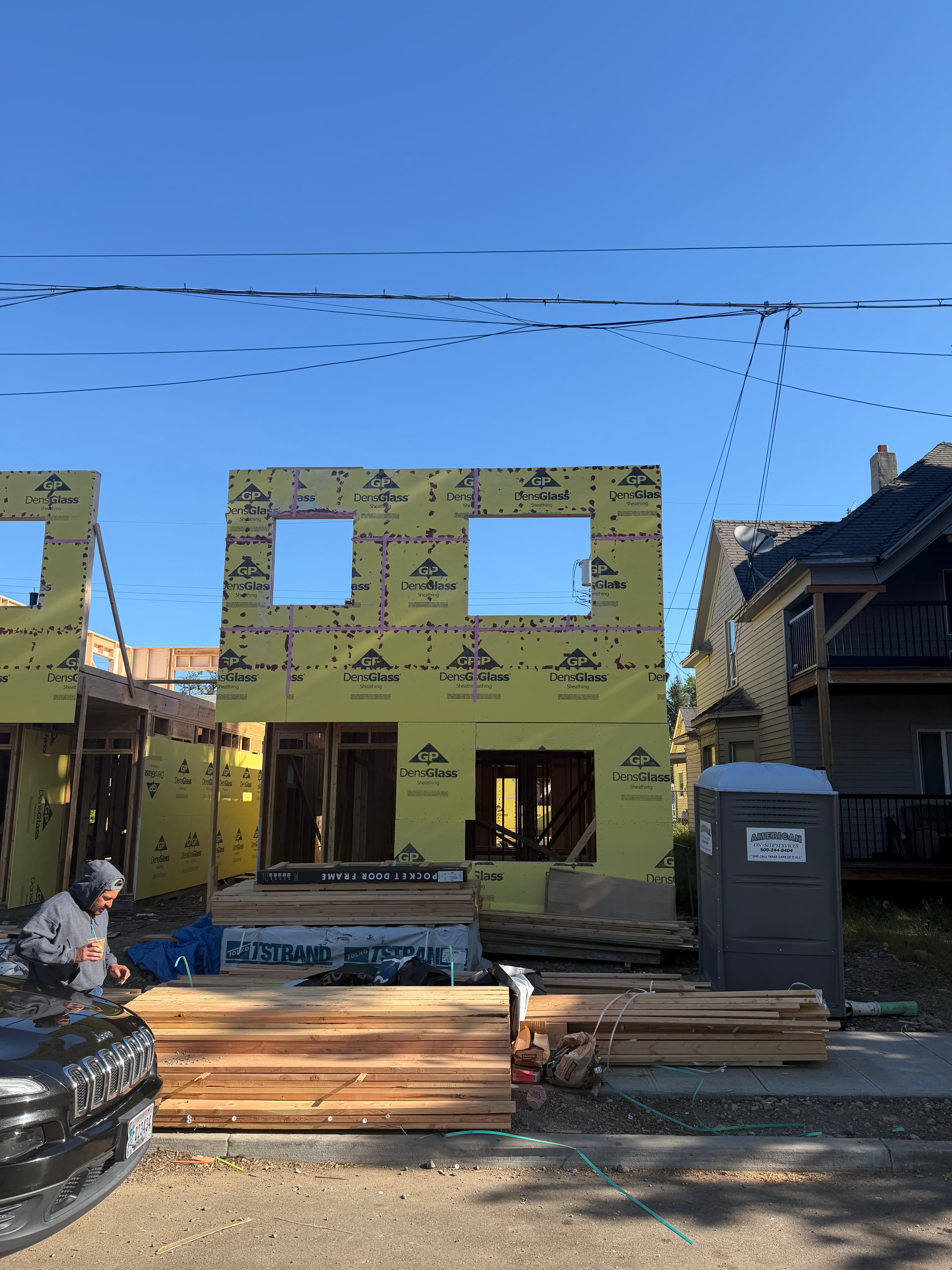 House under construction with yellow sheathing, stacks of lumber, and a portable toilet nearby.