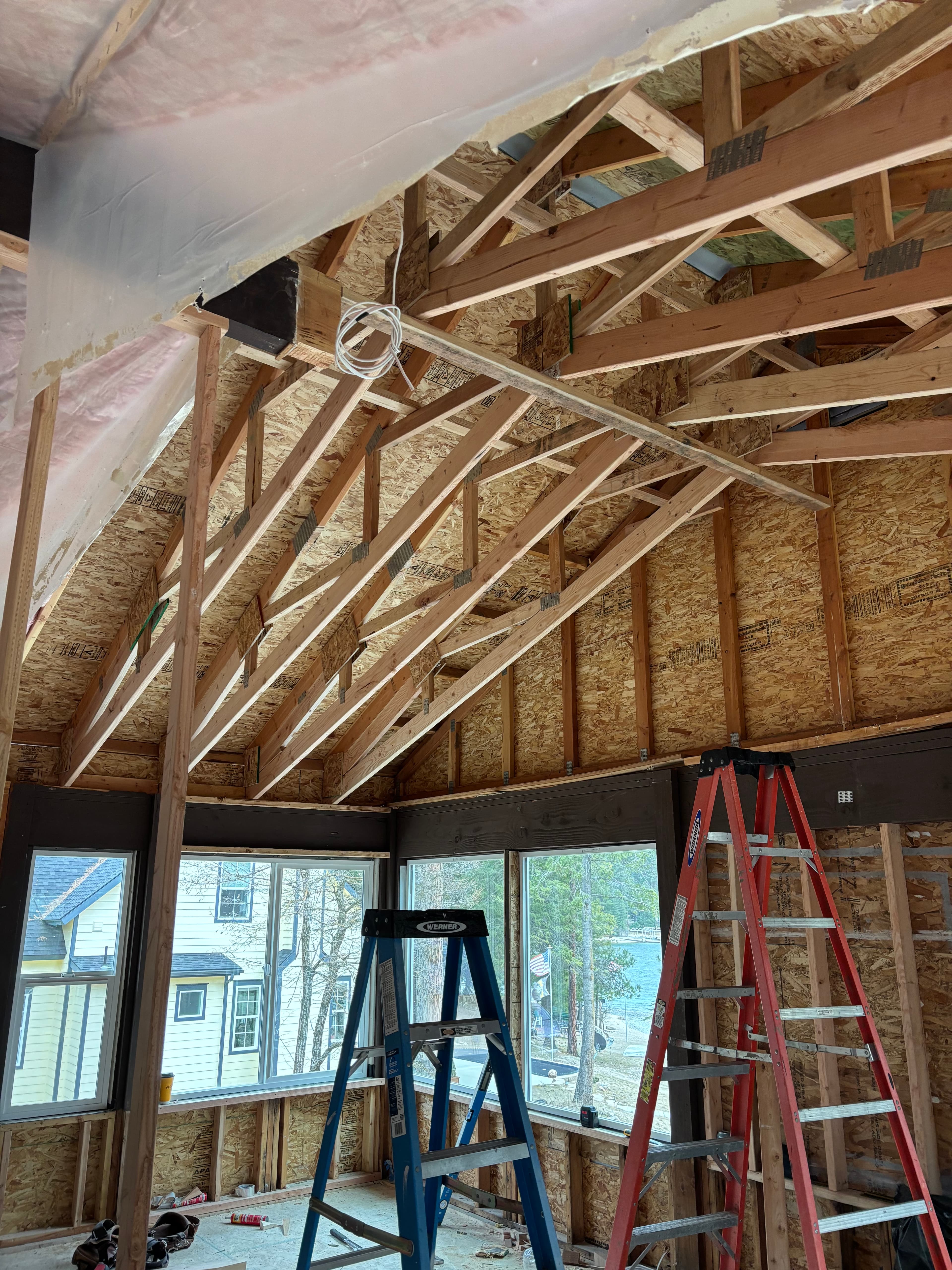 Interior of a house under construction with wooden roof trusses, OSB sheathing, and two ladders.