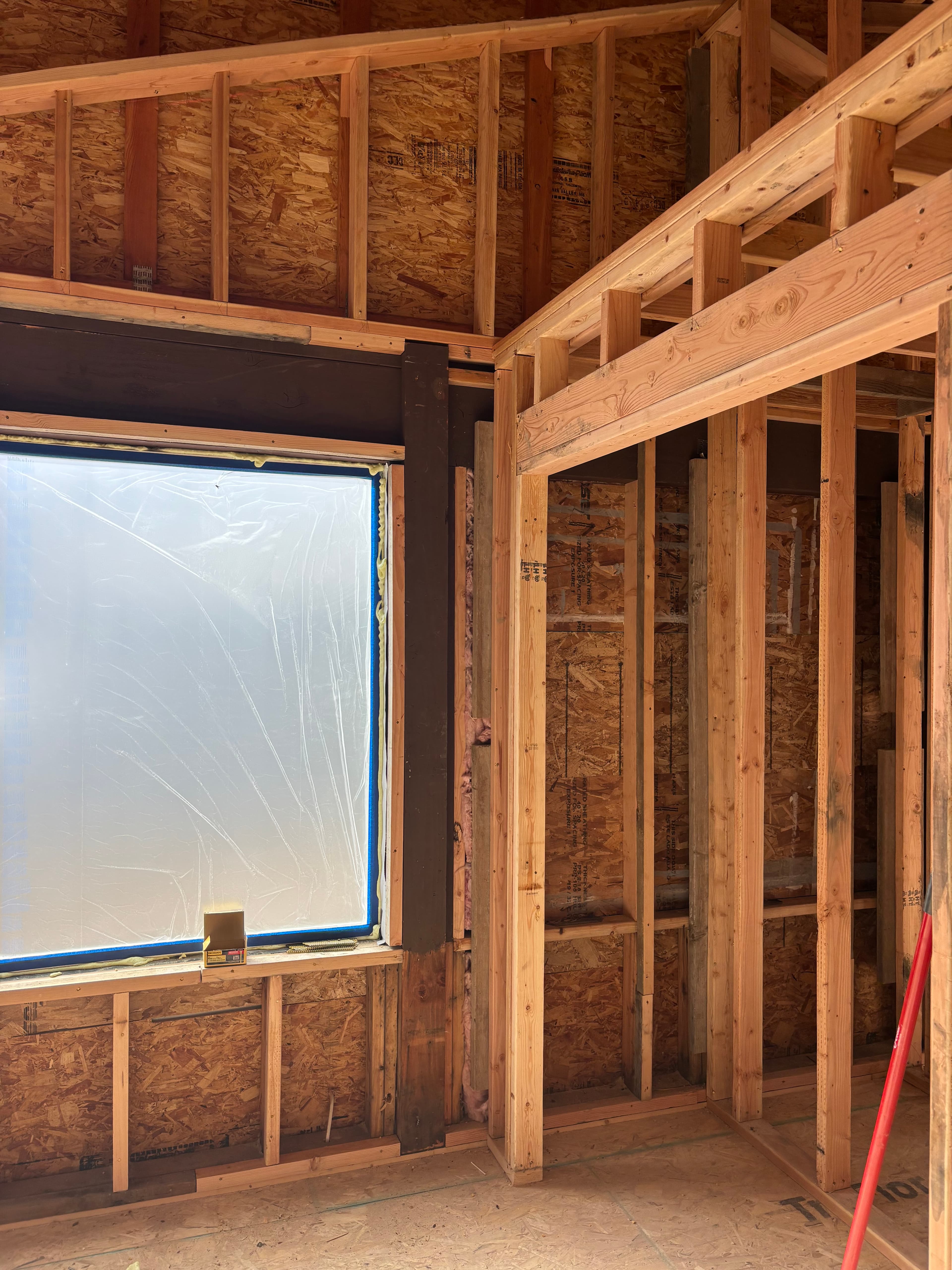 Interior view of wooden house framing and OSB sheathing with a plastic-covered window opening.