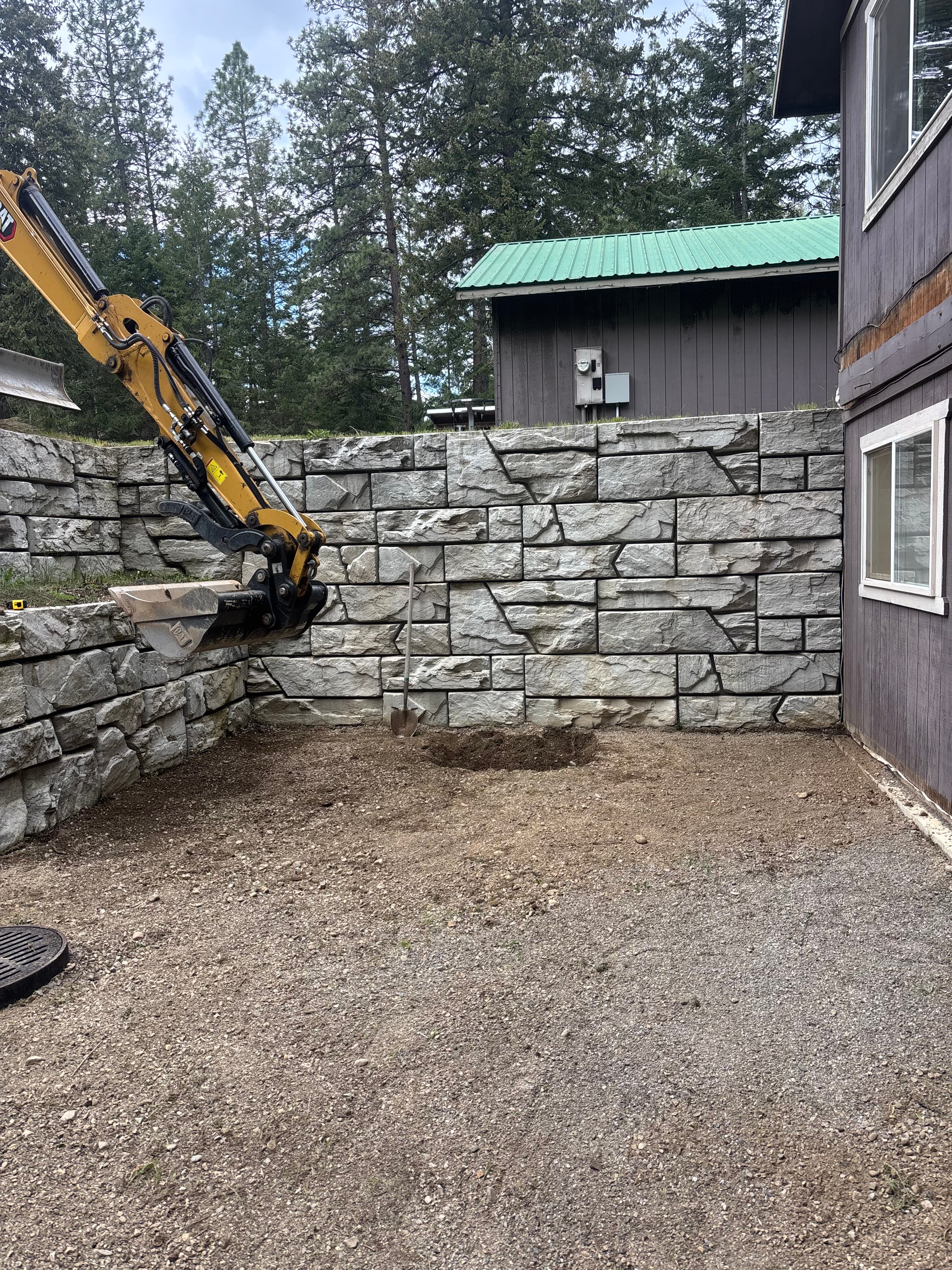 Yellow excavator arm positioned near a large grey stone retaining wall on dirt ground.