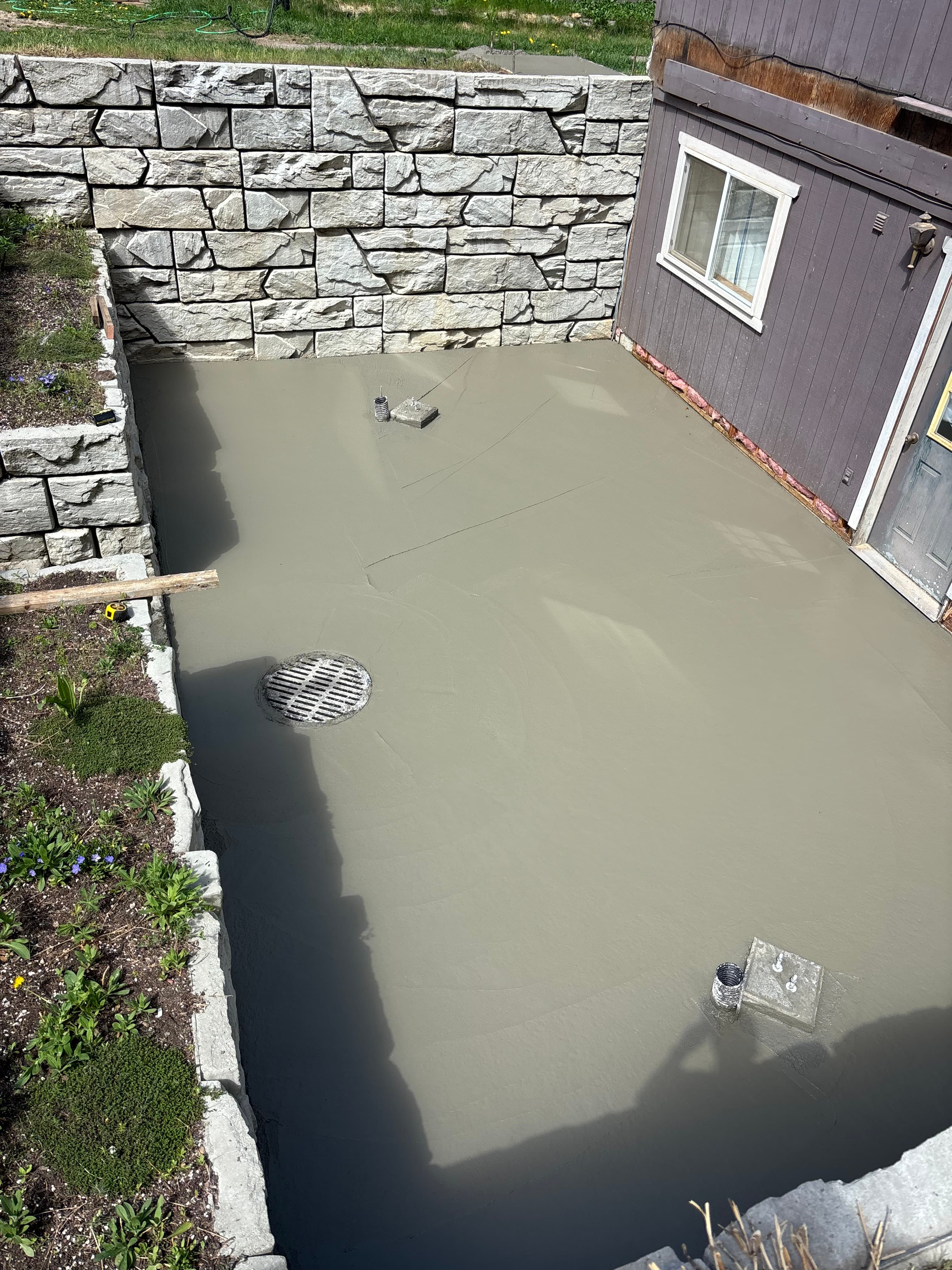 Freshly poured smooth concrete patio with a drain, bordered by a stone retaining wall.